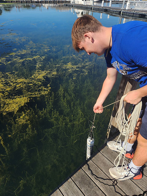 Student standing at the edge of a dock and lowering the Hydrolab data sonde into the water below.